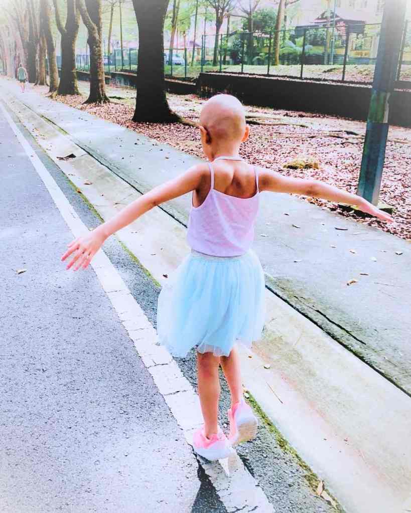 Young girl with a bald head due to chemotherapy as treatment for cancer. She walks along a road with her hands playfully outstretched.